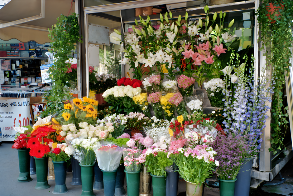 A vibrant display of fresh flowers at a flower shop, featuring white lilies, pink lilies, red gerbera daisies, white roses, pink carnations, purple statice, and various greenery arranged in bouquets a