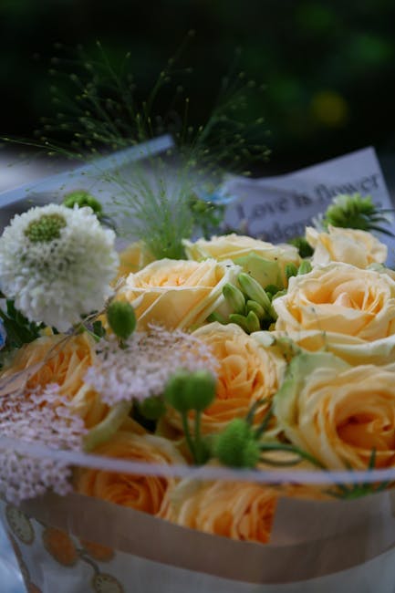 A close-up view of a floral arrangement featuring soft peach roses with delicate, layered petals and fresh green buds. Interspersed among the roses are white pom-pom chrysanthemums and small, fluffy w