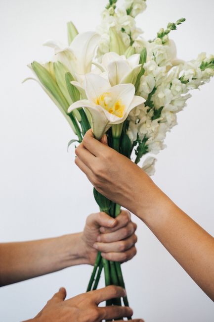 A close-up of two hands holding a bouquet of fresh white flowers, including elegant calla lilies with creamy yellow centers and delicate stock flowers, arranged with lush green stems and leaves. The b