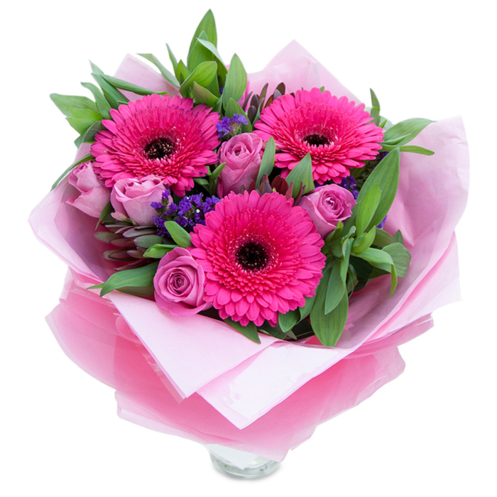 I arranged three striking fuchsia Gerbera daisies as the visual anchors of this bouquet, their broad, layered petals unfurling around pronounced, dark centres that give the composition punctuation and personality. Tucked alongside them are several lavender-pink roses, each bloom neat and gently cocooned, the kind of fresh roses whose outer petals hold the promise of a fuller opening over the next few days. Tiny, vibrant purple statice is peppered throughout, introducing textural lifts and a cooler hue to balance the rosier tones. Lush foliage - the glossy, deep green of salal and the softer, blue-green sweep of eucalyptus - frames every flower, creating depth and a pleasing, garden-like silhouette. The bouquet is thoughtfully wrapped: a subtle pale pink tissue paper forms the inner layer, soft to the touch, while an outer sheet of translucent fuchsia-pink cellophane, slightly crinkled, catches the light and ties the colour story together; the lower folds reveal the rim of a clear glass vase, ready to receive water at once. The fragrance is gentle and comforting, rose notes drifting with a faint, fresh-green undertone from the gerberas. This arrangement feels intimate and optimistic - the sort of gift I would hand over for a birthday on Lordship Lane, to welcome a new neighbour near Goose Green, or to place on a mantel in Dulwich Village; it's designed to convey affection and brighten a room with confident, feminine colour and considered craftsmanship.